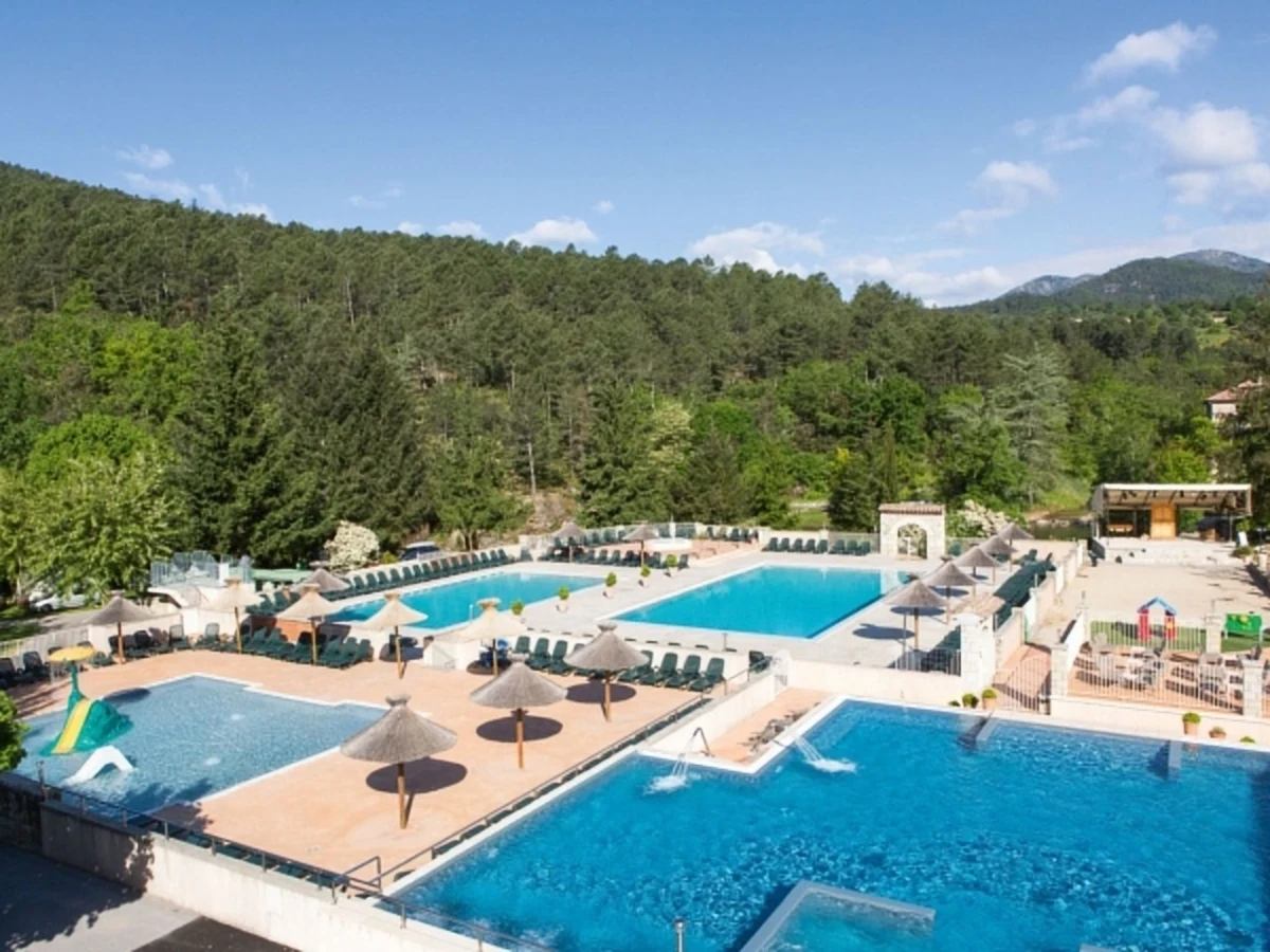 At Les Ranchisses, a spacious swimming pool area features sun loungers and umbrellas, encircled by verdant, wooded hills.