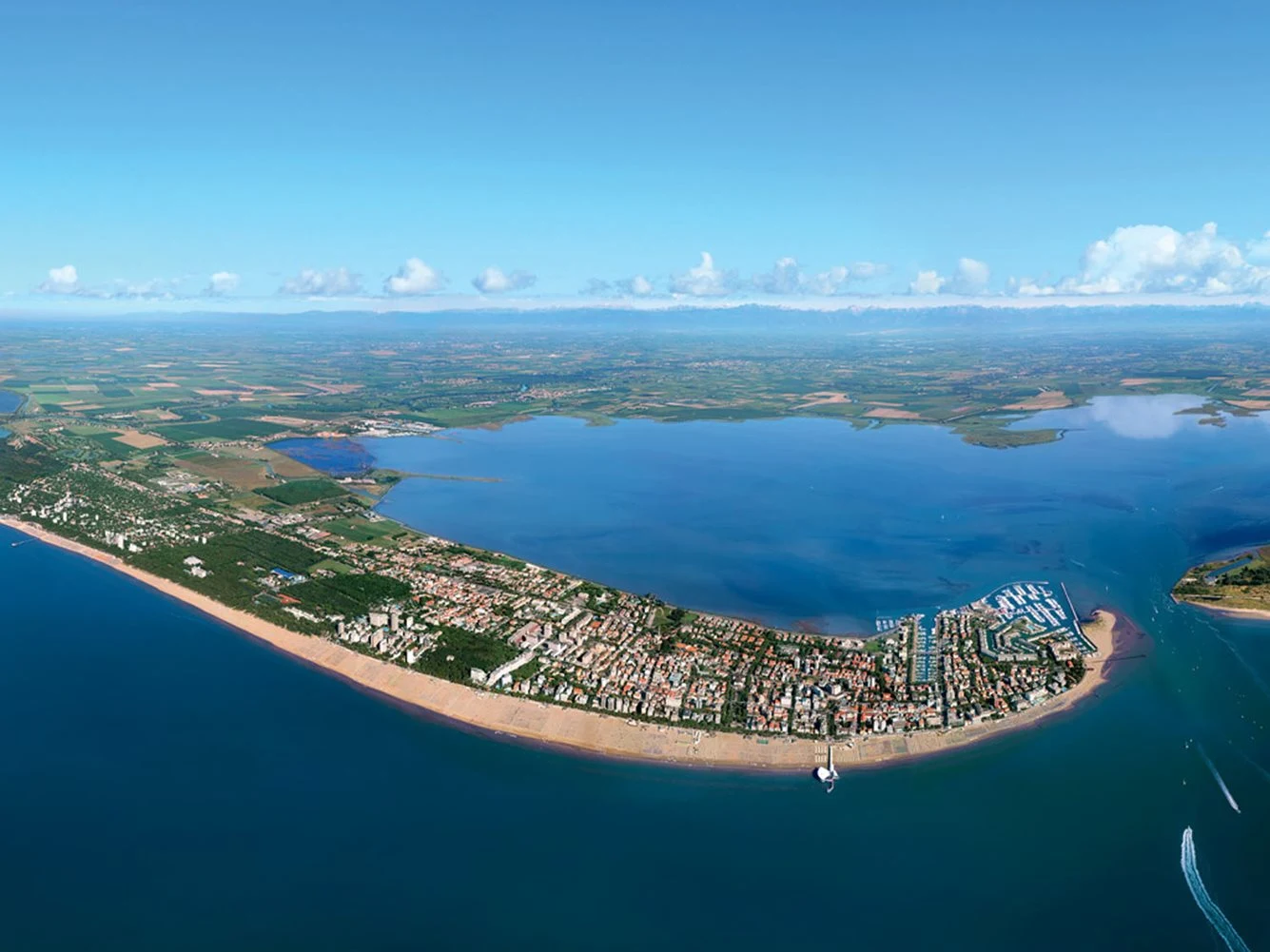 At Pino Mare, a bird's-eye view reveals a slender seaside village nestled between a sandy shore and a vast inland lagoon, encircled by lush fields.