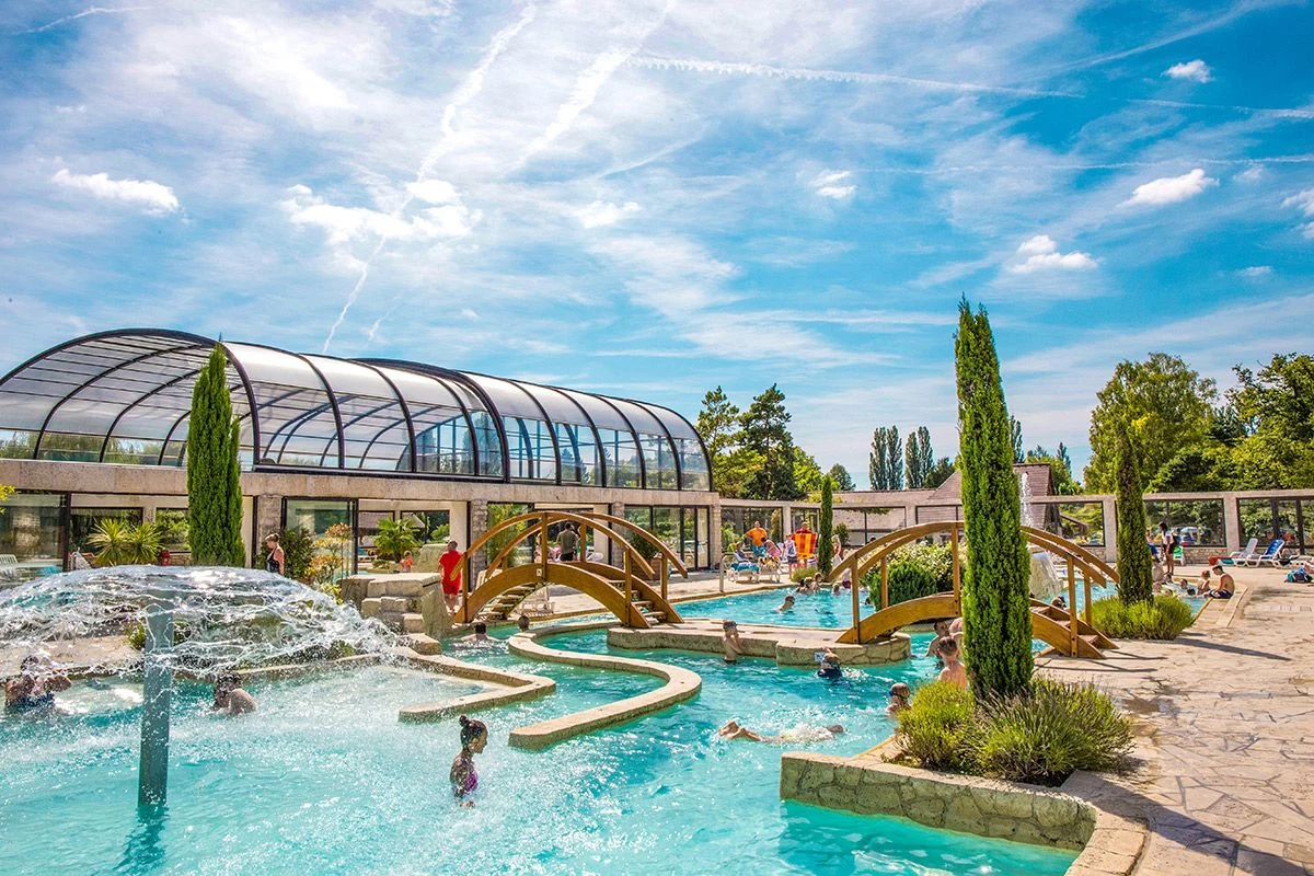 At Marvilla Parks La Croix du Vieux Pont, a swimming area with walkways and water features is encircled by individuals relishing the water beneath a bright sky.