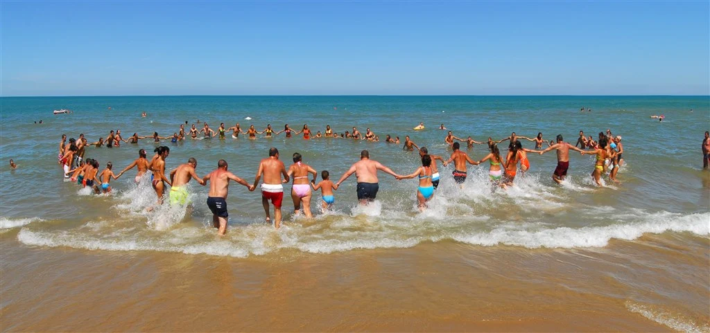 At Stella del Sud, individuals grasping each other's hands create a large ring while splashing in the shallow waters of the ocean on a sunny beach.