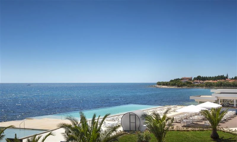 At Aminess Sirena, the swimming pool provides a view of the ocean, encircled by sun loungers and palm trees, with a backdrop of clear blue skies.