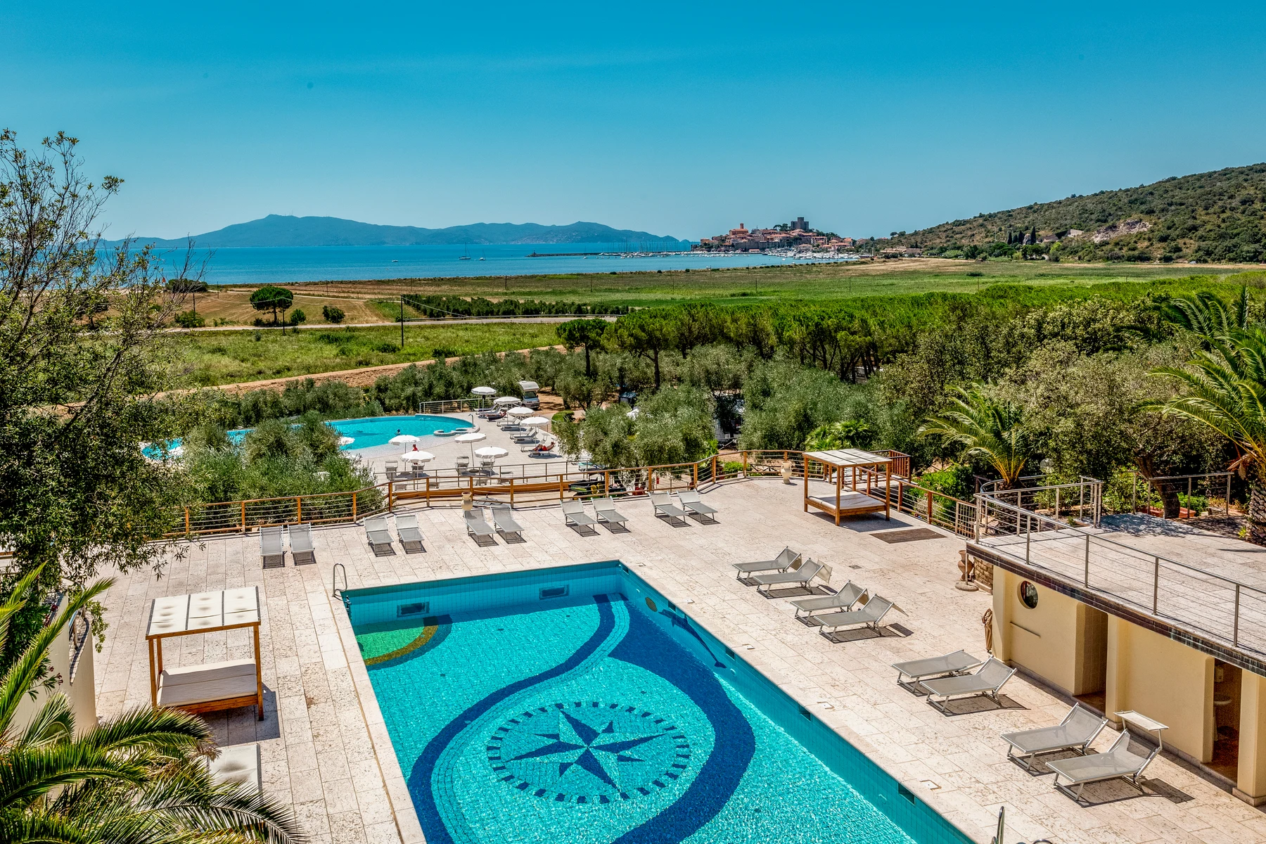 At Gitavillage Talamone, a swimming pool encircled by loungers is visible, with a distant coastal town and verdant foliage framing the landscape. The setting is sunny and outdoors.