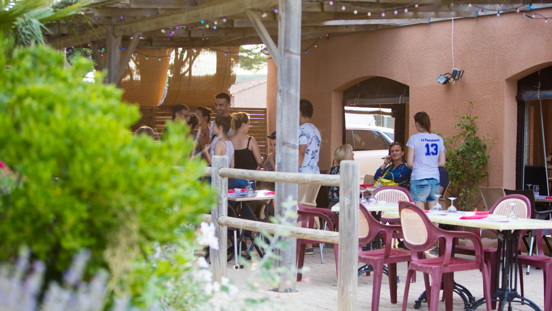 At Pascalounet, individuals are mingling beneath a wooden pergola, encircled by tables and chairs, in a laid-back outdoor atmosphere.