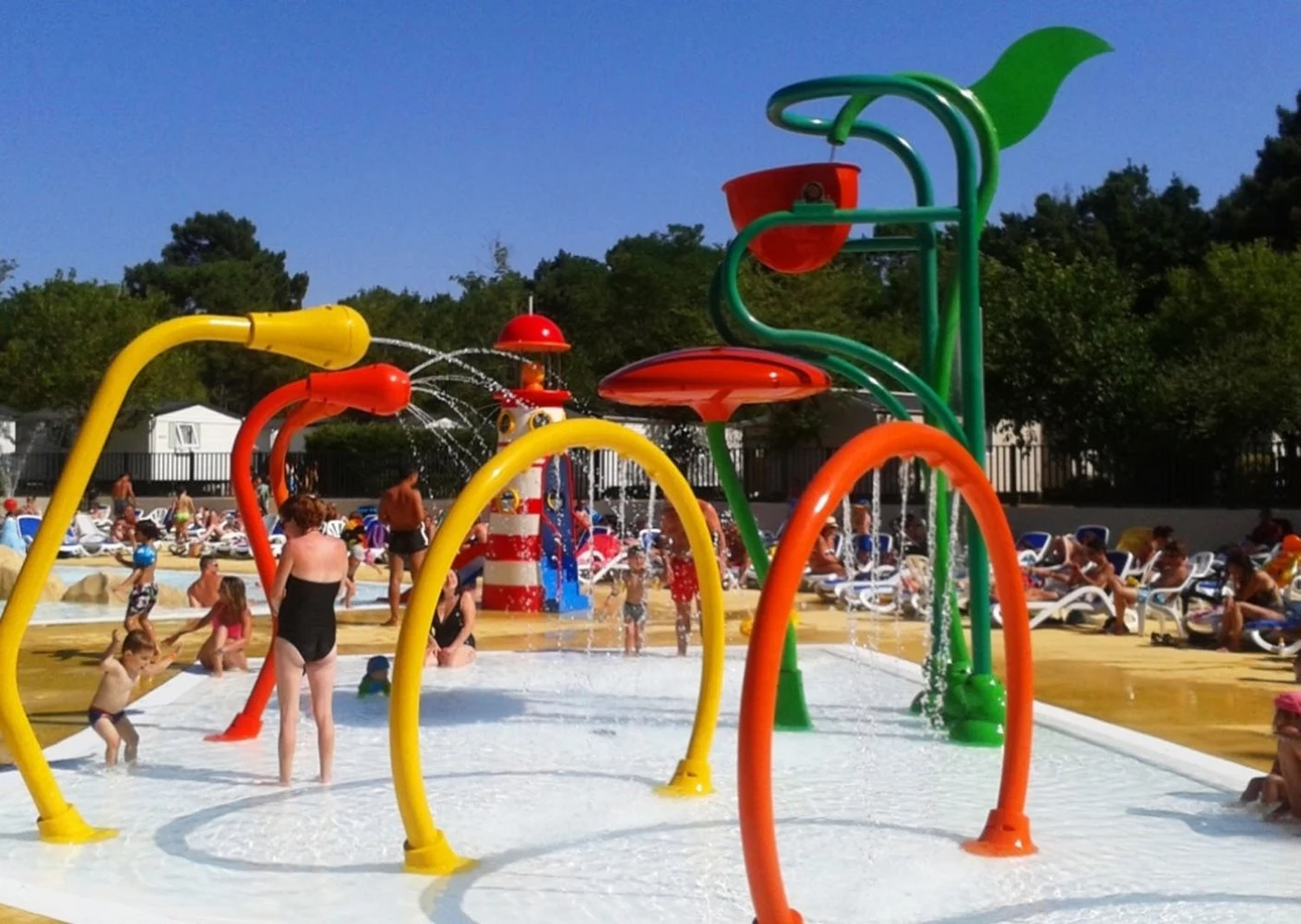 At Palmyre Loisirs, kids are enjoying themselves beneath vibrant water sprays at a bustling outdoor splash pad, encircled by sunbathers and trees.