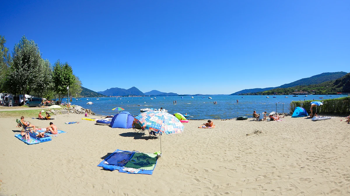 At Orchidea, parasols and sunbathers unwind on a sandy shore next to a tranquil lake, with far-off mountains and scattered vessels.