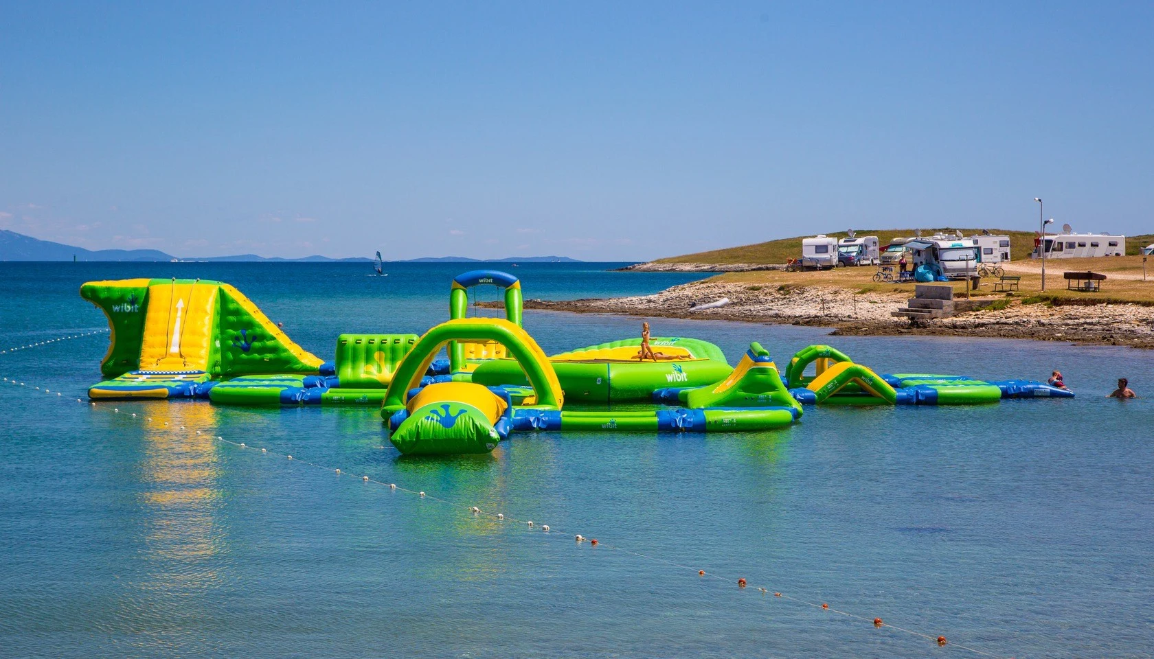 At Stupice, a buoyant water amusement park drifts on a tranquil sea, with campers and recreational vehicles visible on the far shore.