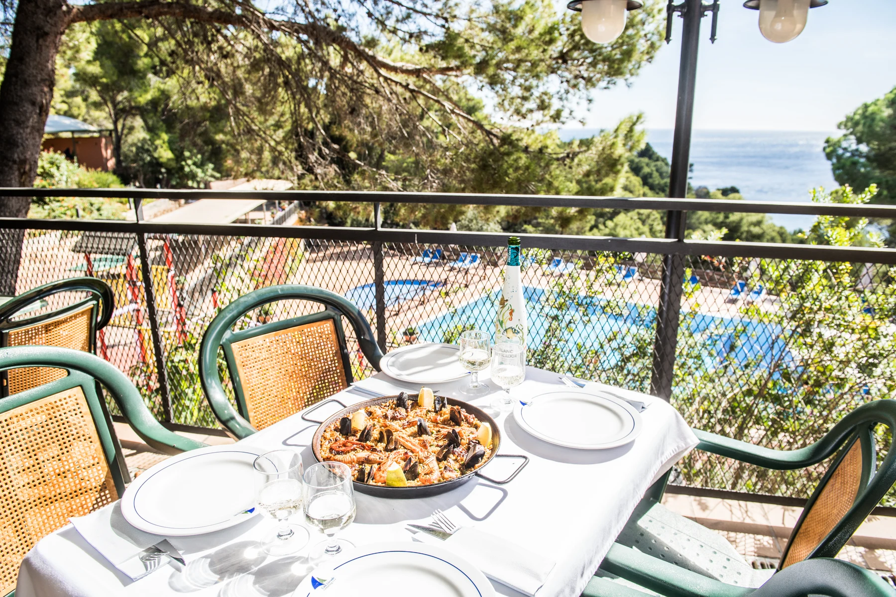 At Cala Llevado, a paella is set on a table with dishes and wine, providing a view of a pool and the sea, encircled by trees.