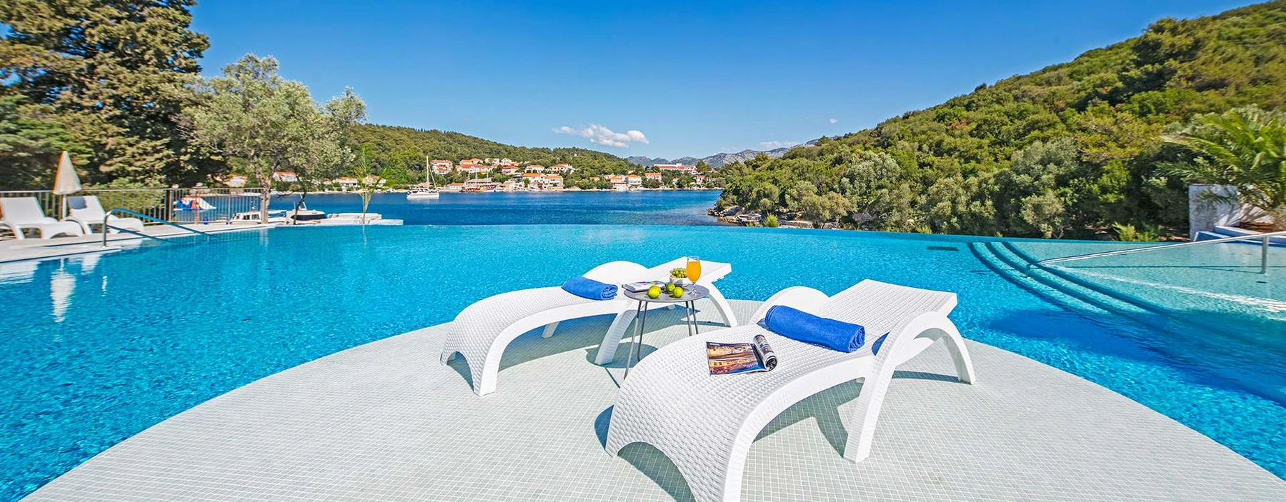 At Aminess Port 9, two ivory lounge chairs face an endless pool beside a tranquil lake, encircled by verdant hills.