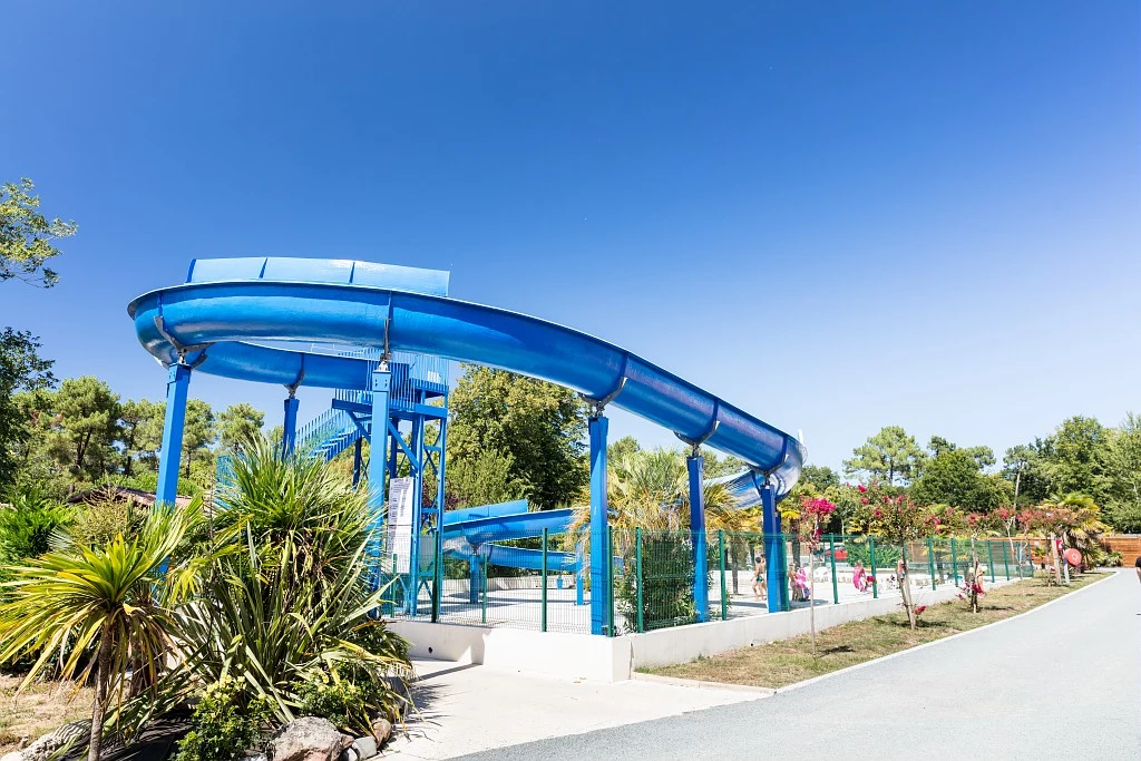 At Marvilla Parks La Clairière, a blue water slide features individuals strolling beneath it in a sunny outdoor park.