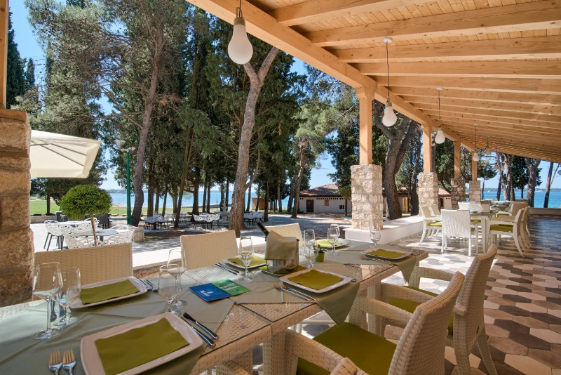 At Pineta, dining tables are arranged beneath a wooden shelter, providing a view of a wooded area with a glimpse of the ocean.