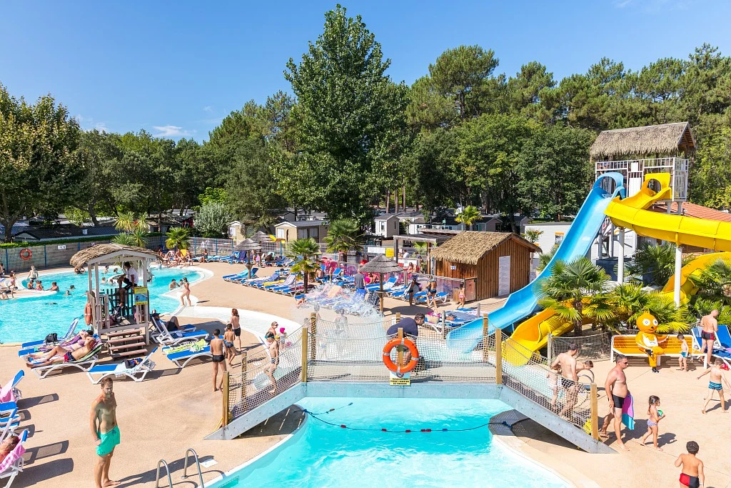 At Lou Pignada, individuals are relishing swimming and sunbathing by a pool featuring water slides, encircled by trees and cabins.