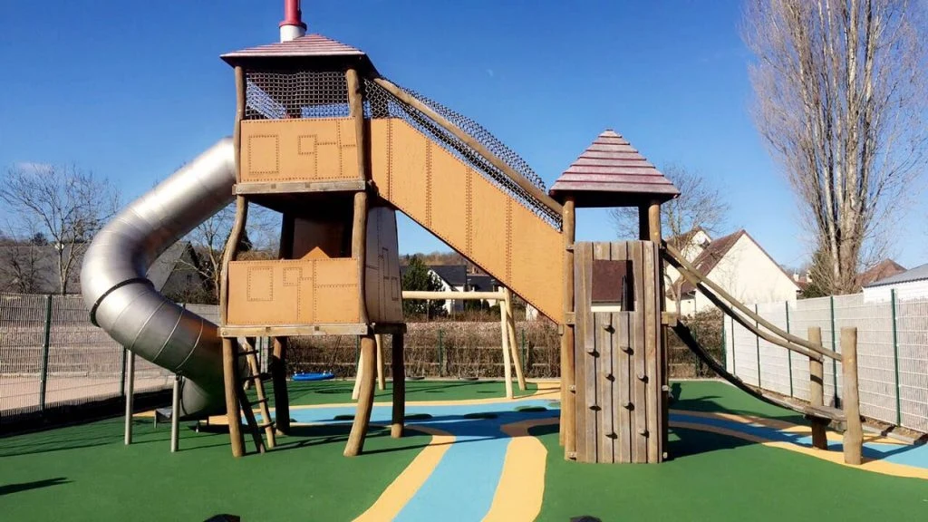 At La Vallée de Deauville, there is a children's play structure featuring slides and climbing zones on a vibrant rubber surface, encircled by trees and buildings.