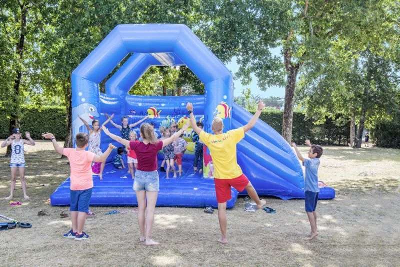 At Domaine d'Oléron, kids are enjoying themselves on a blue inflatable castle, overseen by an adult, amidst trees and greenery.