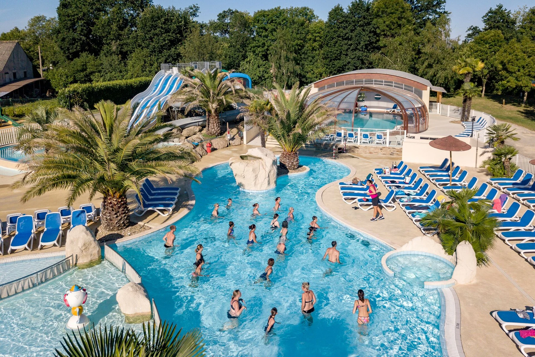At Les 2 Fontaines, individuals are working out in a spacious outdoor swimming pool encircled by palm trees and deck chairs.