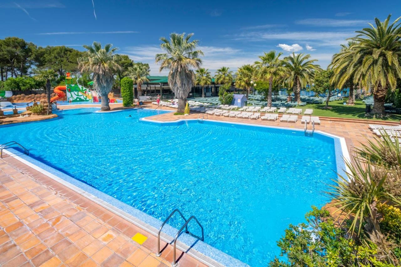 At Solmar, a spacious swimming pool features sun loungers, encircled by palm trees and a clear blue sky.