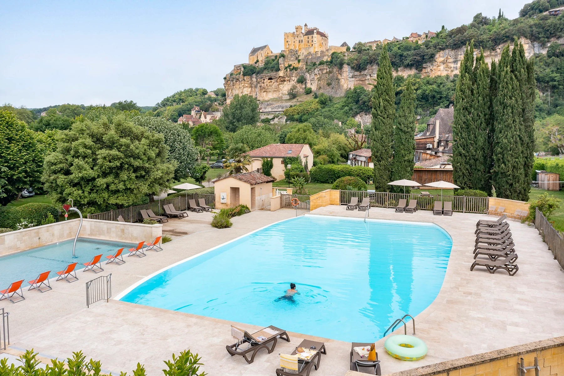 At Le Capeyrou, an individual is swimming in a spacious outdoor pool, encircled by sun loungers and lush vegetation, with a striking cliff and castle in the backdrop.