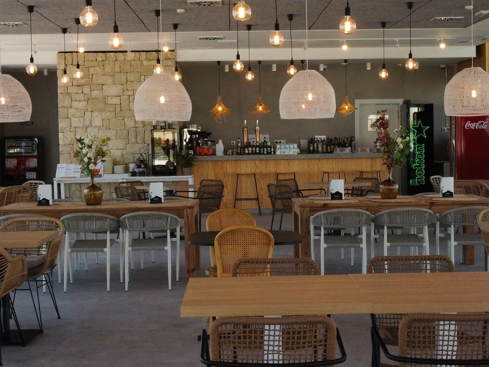 At La Media Legua at the front, there is a seating area featuring wicker chairs positioned around wooden tables beneath suspended lights, adjacent to a stone-walled bar in a café.