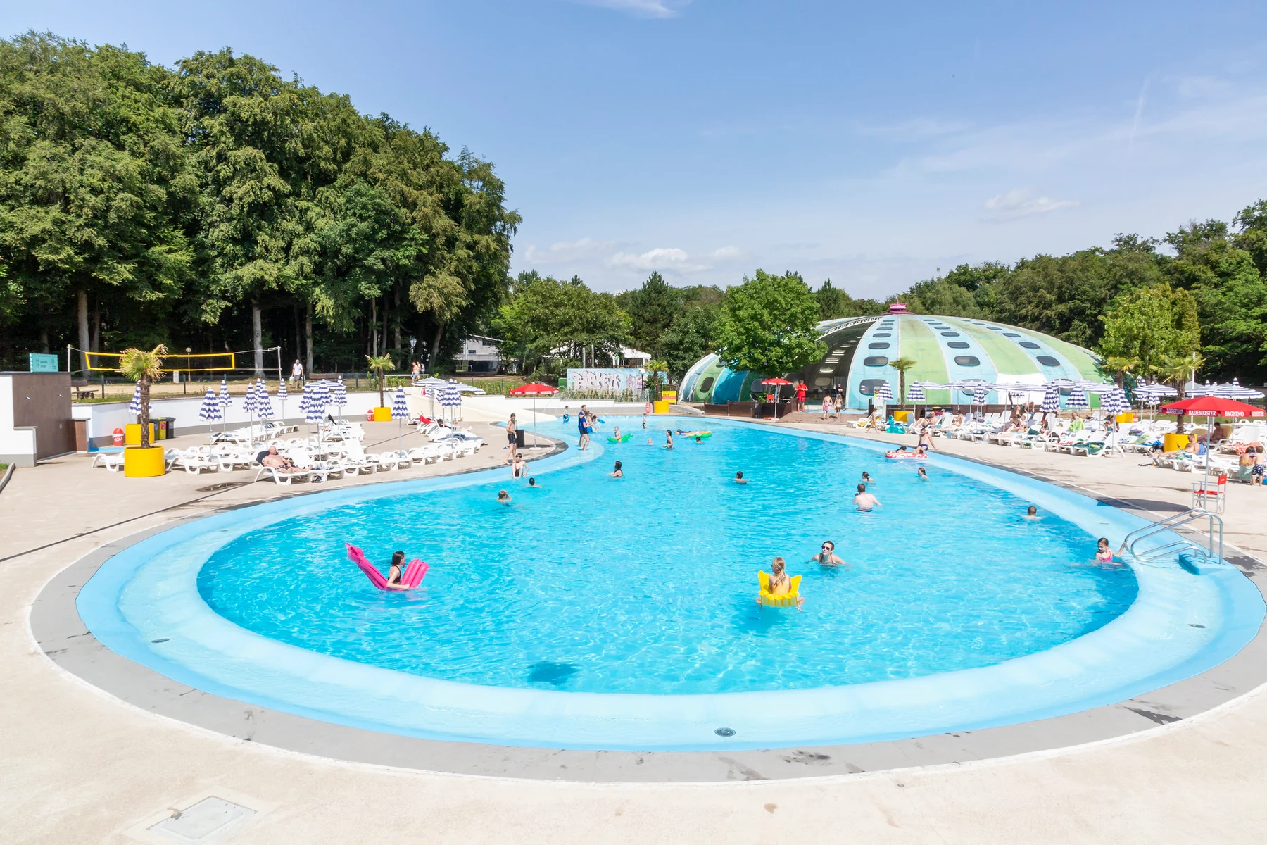 At Birkelt Village, a swimming pool features individuals enjoying games and unwinding, encircled by loungers and parasols, adjacent to a green dome structure in a sunny, wooded area.
