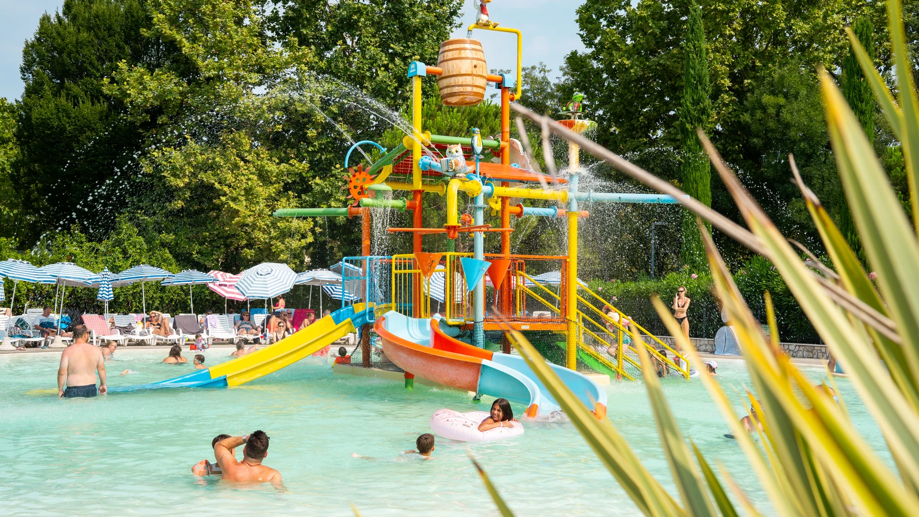 At Bella Italia, a vibrant water play area is spraying water, encircled by children enjoying themselves in a swimming pool, with trees and loungers in the vicinity.