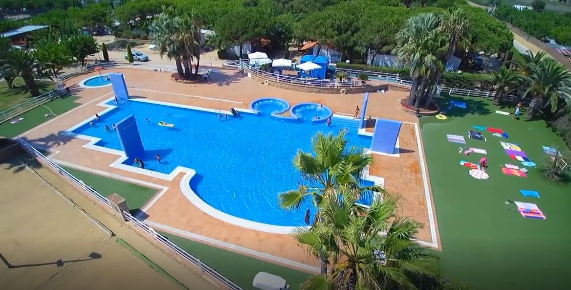 At El Pla de Mar, a spacious swimming pool is encircled by sunbathers and palm trees, with a grassy area featuring towels and umbrellas.