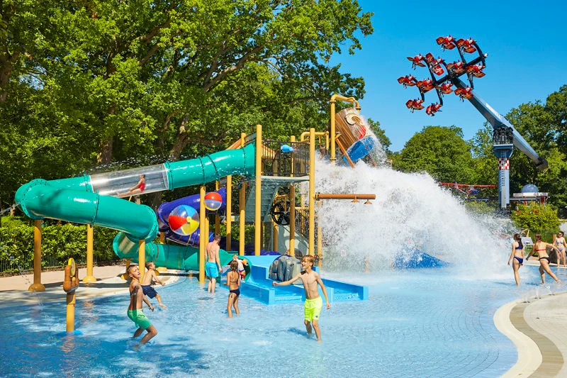 At Duinrell, there is a water play zone featuring slides and splash attractions; youngsters are enjoying themselves in the shallow pool, which is encircled by trees and amusement rides.