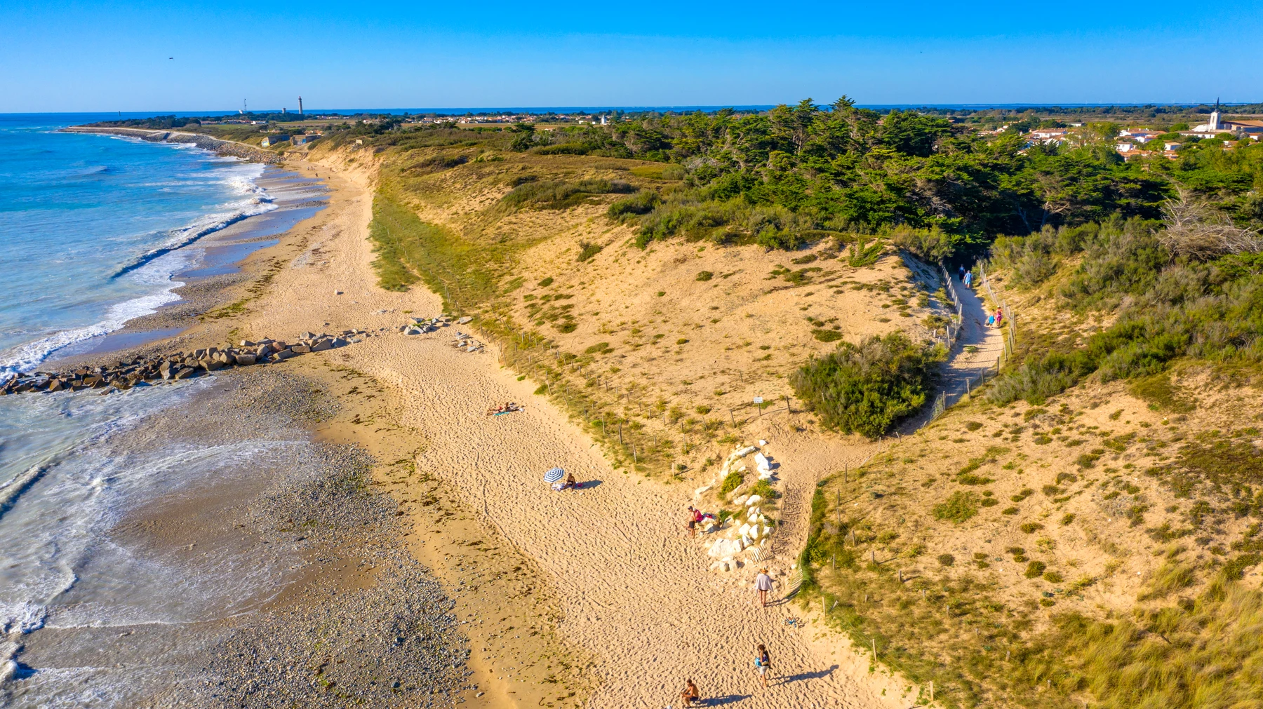 At Marvilla Parks La Côte Sauvage, individuals stroll along a sandy trail leading to a seaside beach, accompanied by waves and lush vegetation lining the coast.
