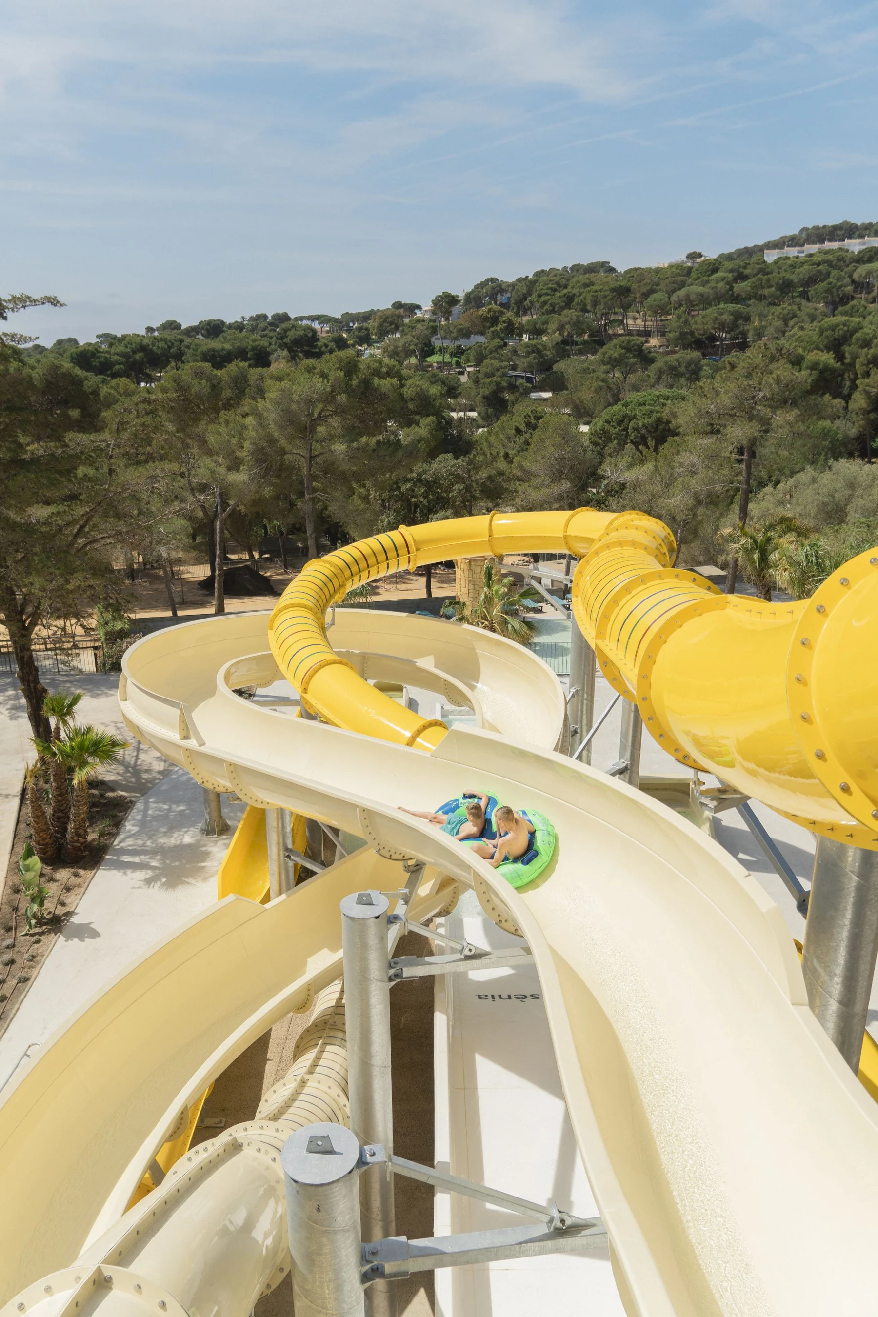 At Internacional de Calonge, two individuals are navigating a winding water slide on an inflatable raft, encircled by vibrant trees.