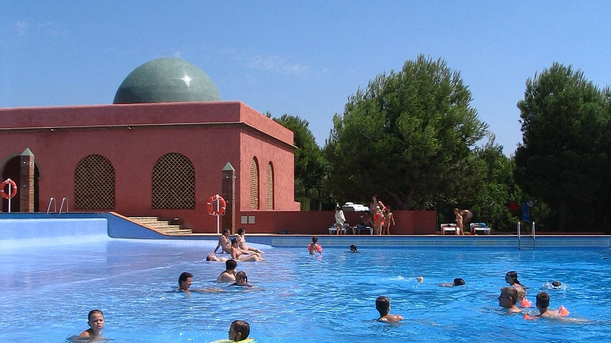 At El Templo del Sol, individuals are swimming in a vast azure pool adjacent to a crimson structure topped with a verdant dome, encircled by trees.