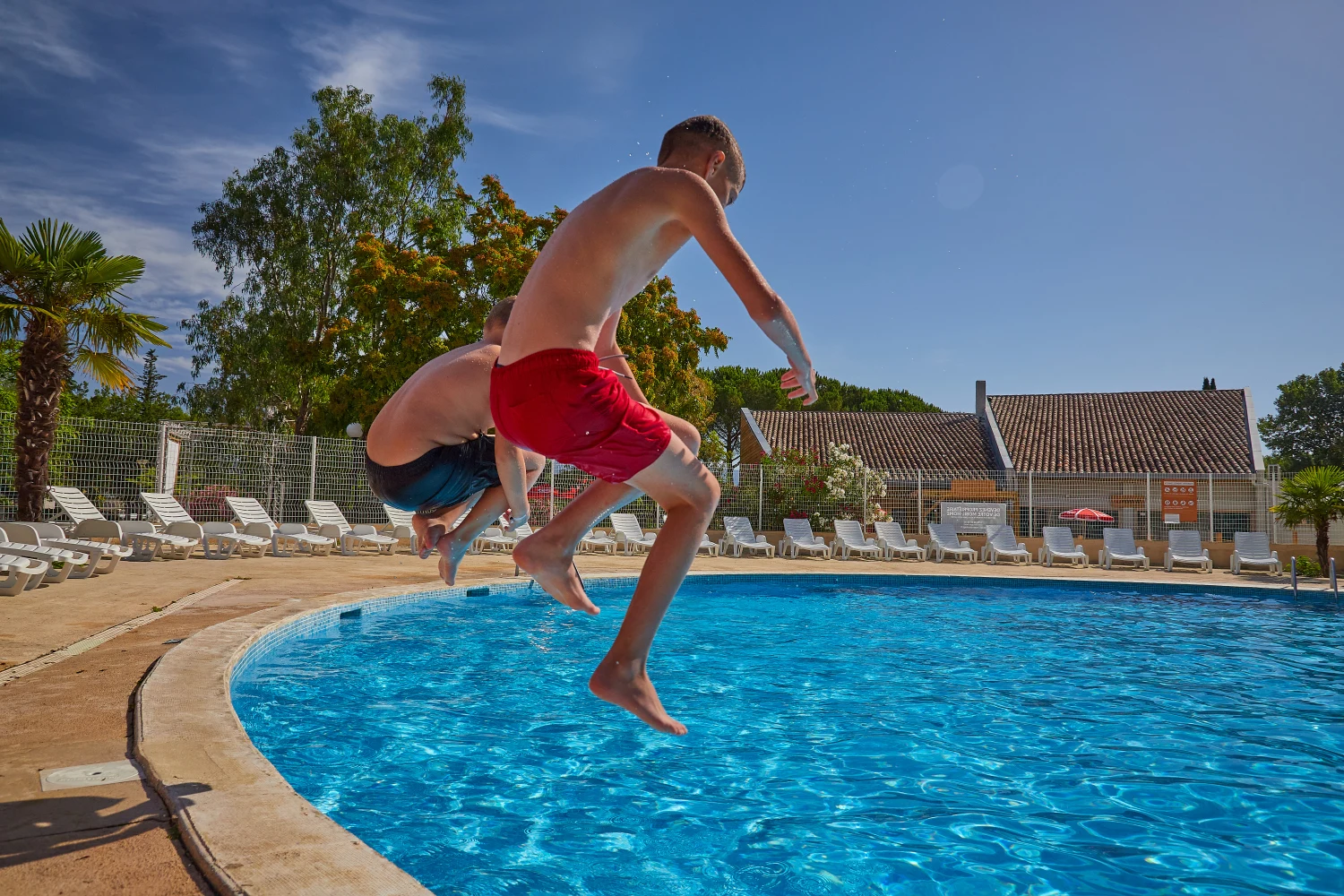 At Domaine des Iscles, two individuals leap into a swimming pool, encircled by deck chairs and trees.