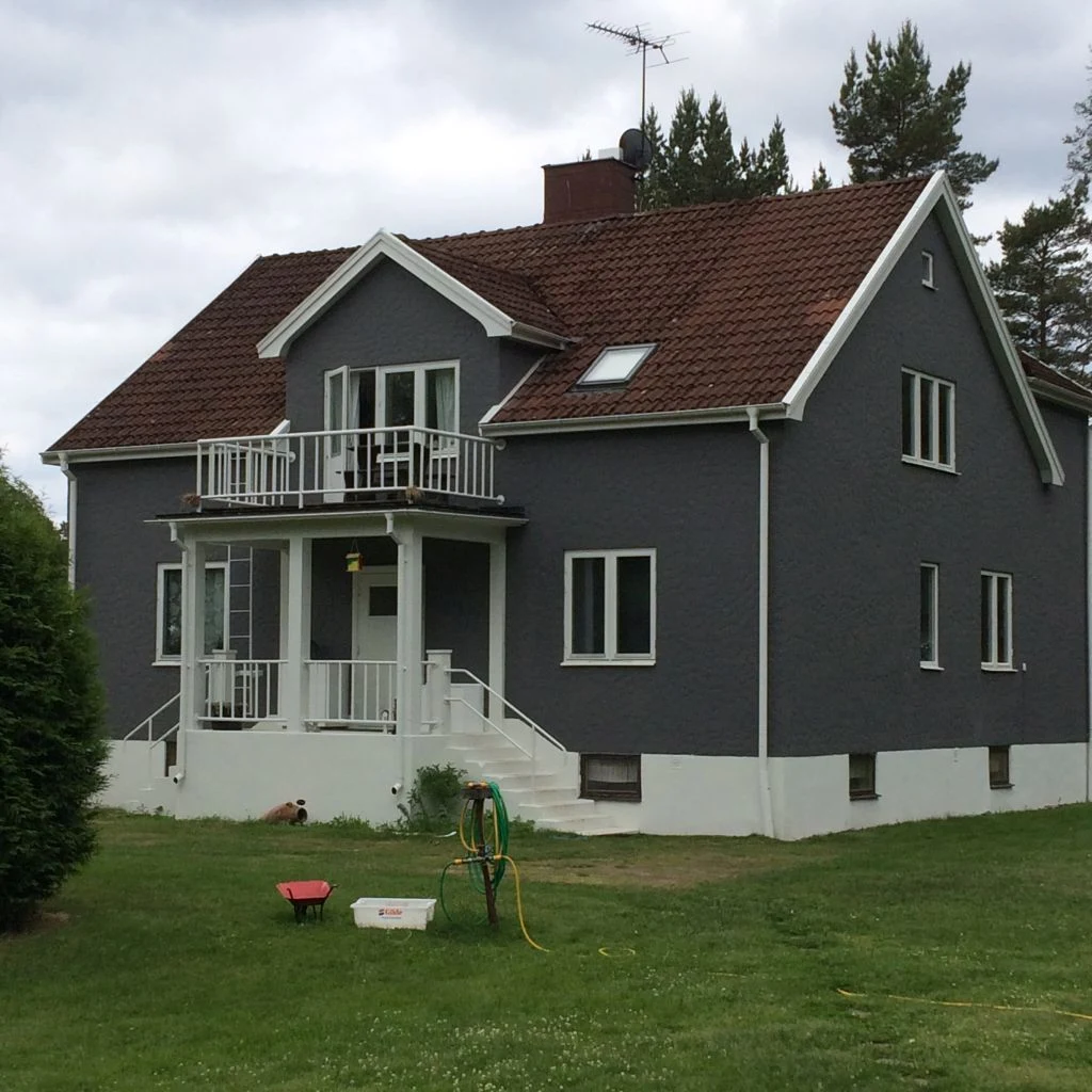At Smaland Miniglamping, a gray two-story home with a crimson roof is located on a verdant lawn, encircled by trees. A wheelbarrow and hose are positioned in the yard.