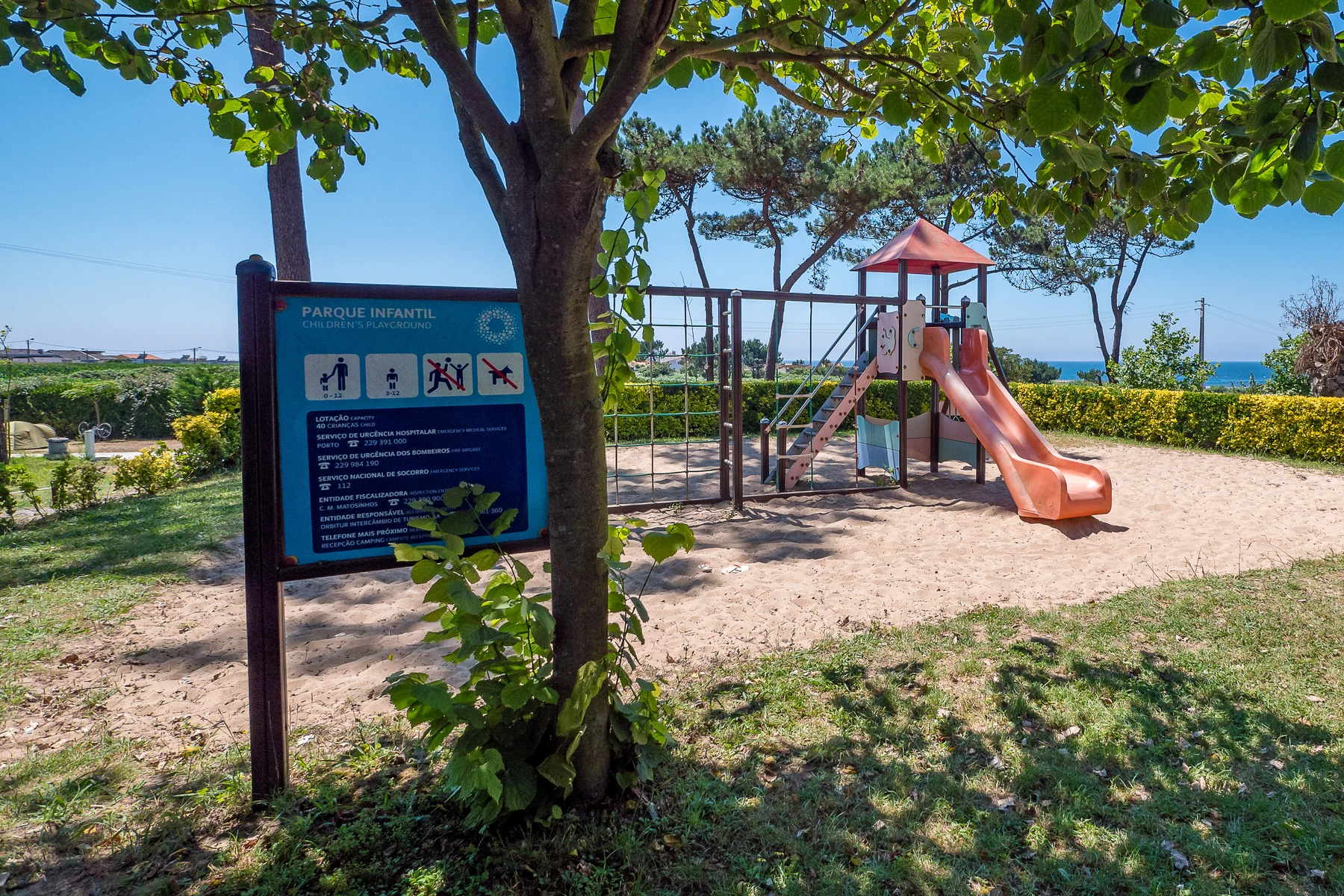 At Angeiras, playground apparatus remains idle on sandy terrain, encircled by lush vegetation and a signboard.