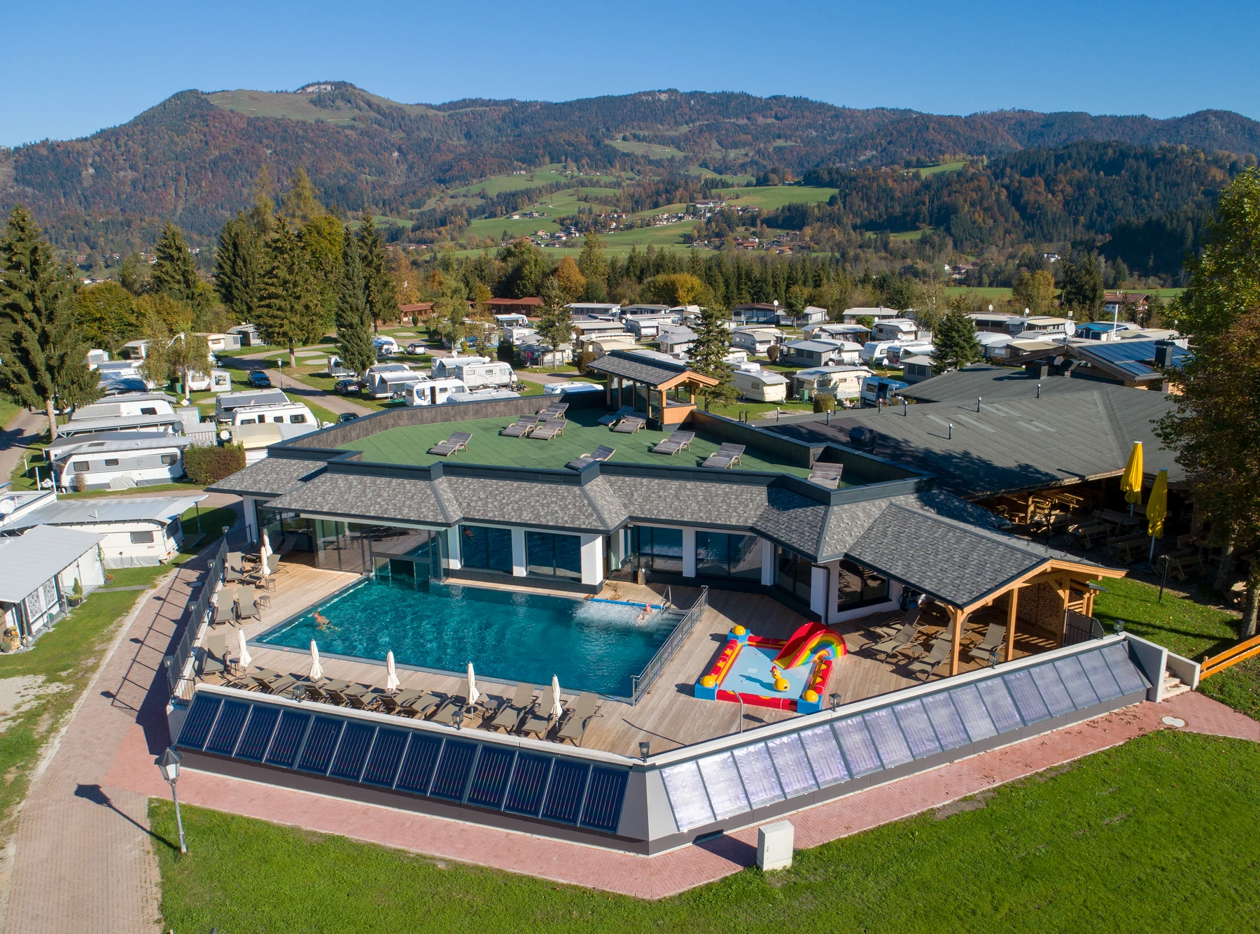 At Eurocamp Wilder Kaiser, a swimming pool is located beside a sizable structure, encircled by many caravans and bordered by verdant trees and a hilly terrain.