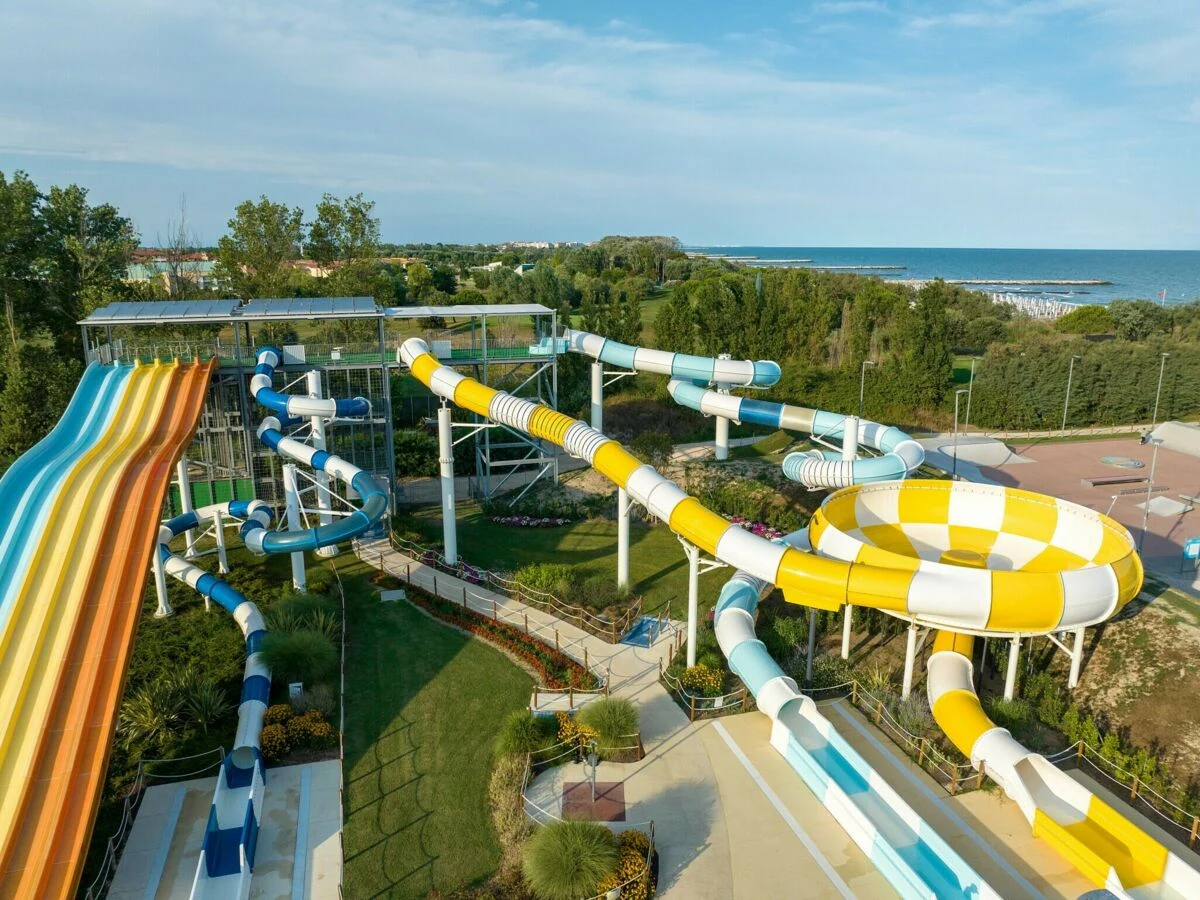 At Pra delle Torri, vibrant water slides spiral and curve, encircled by lush vegetation, with a view of a far-off coastline.