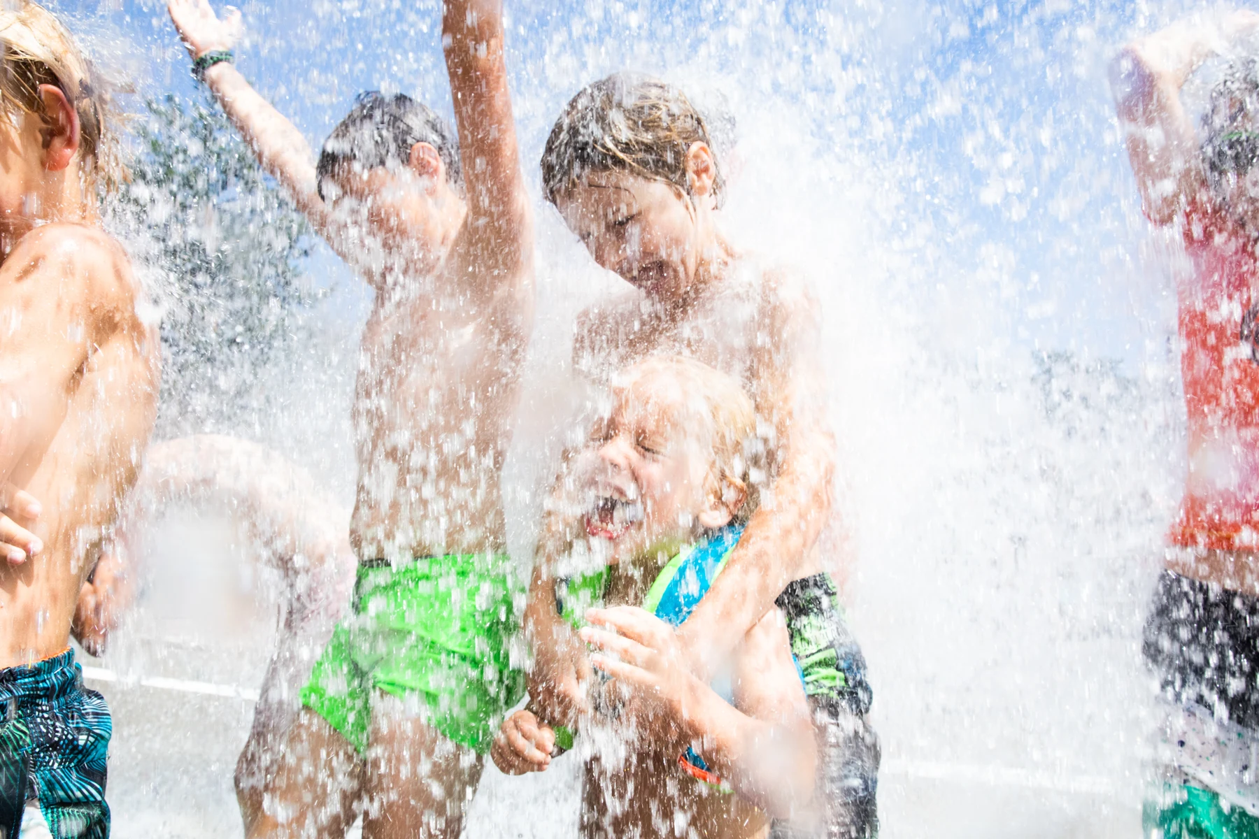 At Het Grote Bos, children frolic happily beneath flowing water, arms lifted, in a vibrant outdoor environment.