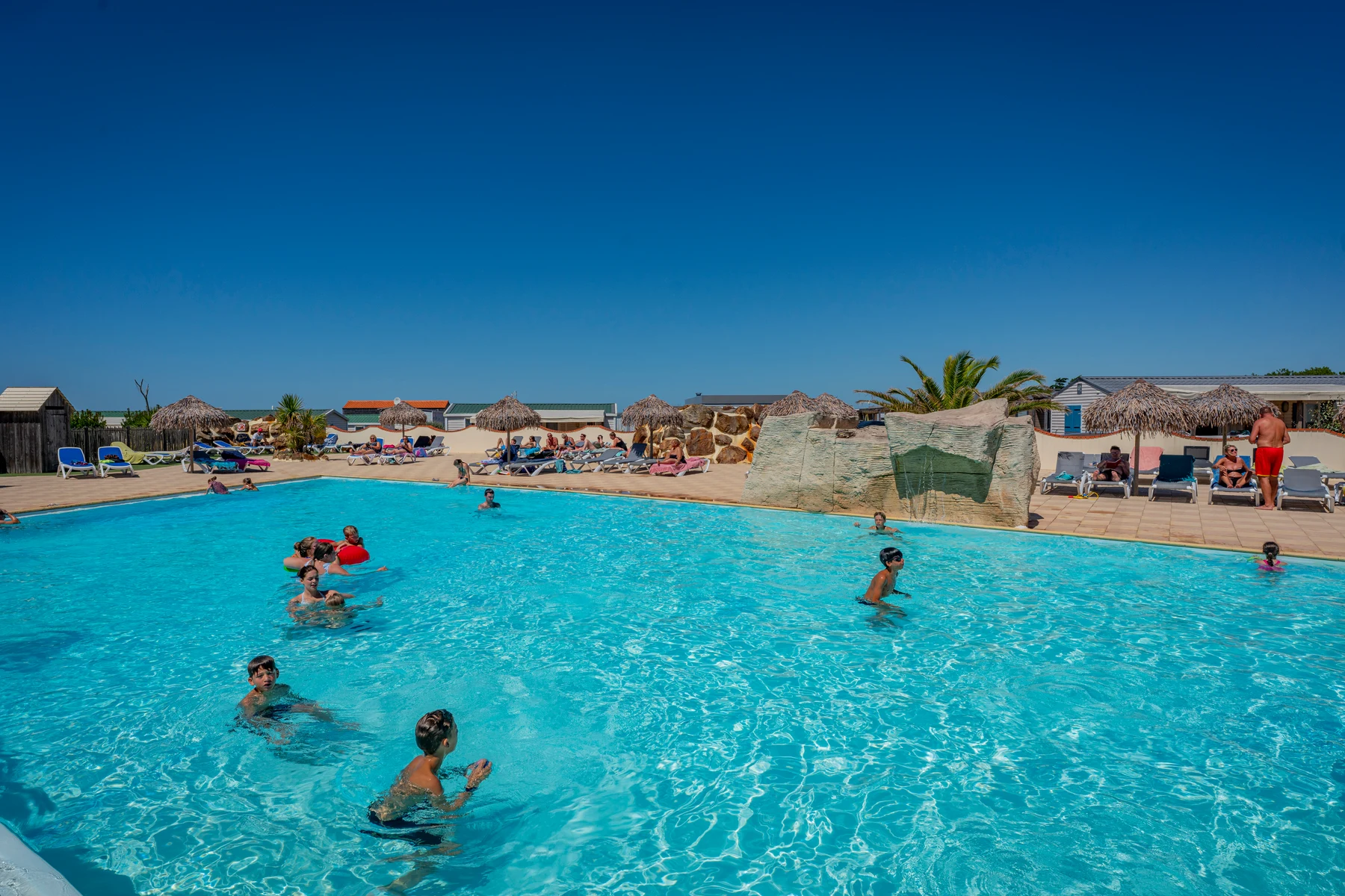 At La Dune des Sables, individuals are swimming in a crystal-clear blue pool, encircled by sun loungers and straw huts beneath a bright sky.