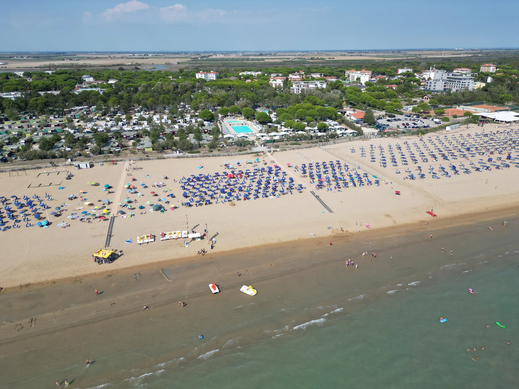 At Lido - Bibione, individuals are unwinding beneath vibrant umbrellas on a sandy shore, next to a swimming pool and a camping area.