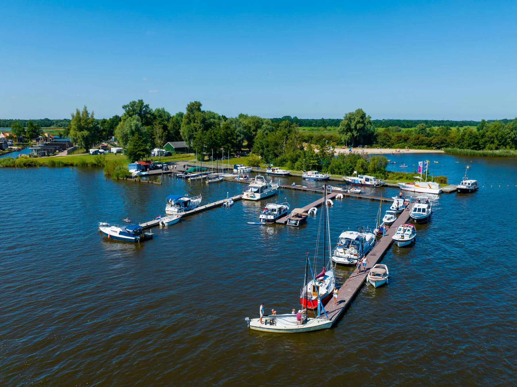 At Vakantiepark Bergumermeer, vessels are moored along timber docks, individuals are strolling, and the area is encircled by trees and grassy spaces.