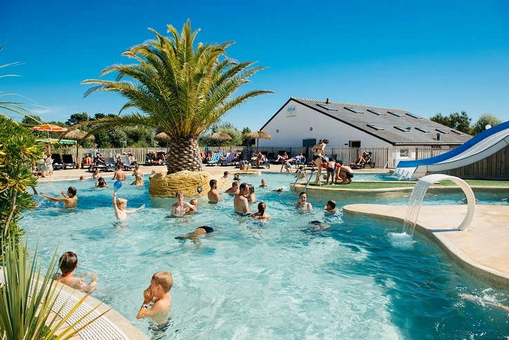 At Les Iles, individuals are swimming and enjoying themselves in a pool, encircled by lounge chairs and a waterslide, with a nearby building.