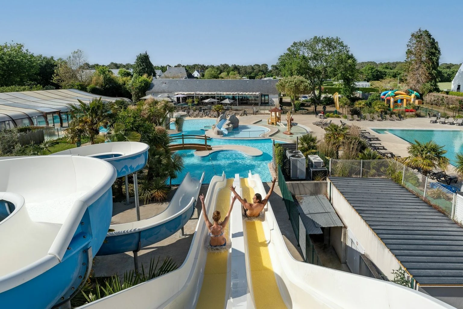 At Carnac, two individuals glide down water slides into a swimming pool, encircled by lush vegetation and deck chairs.