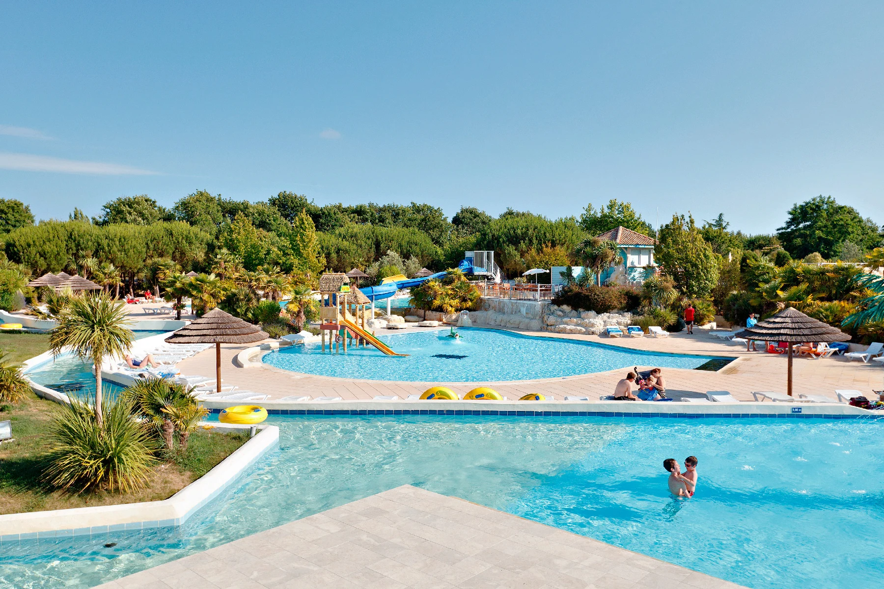 At Sequoia Parc, individuals swim and engage in activities within a spacious outdoor pool facility encircled by lush vegetation and shaded relaxation zones.