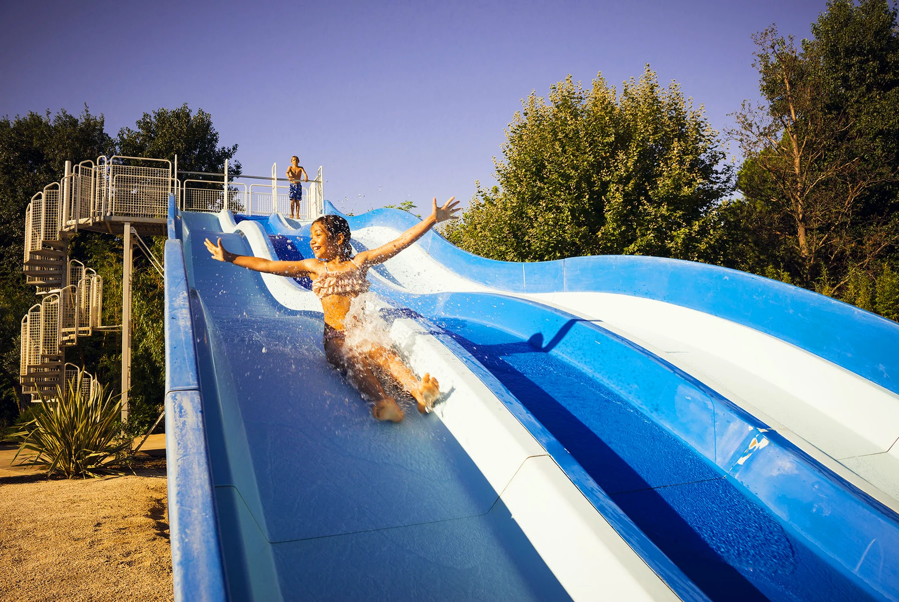 At Le Dauphin, a child glides down a water slide with arms extended, giggling among the trees and under the clear skies.