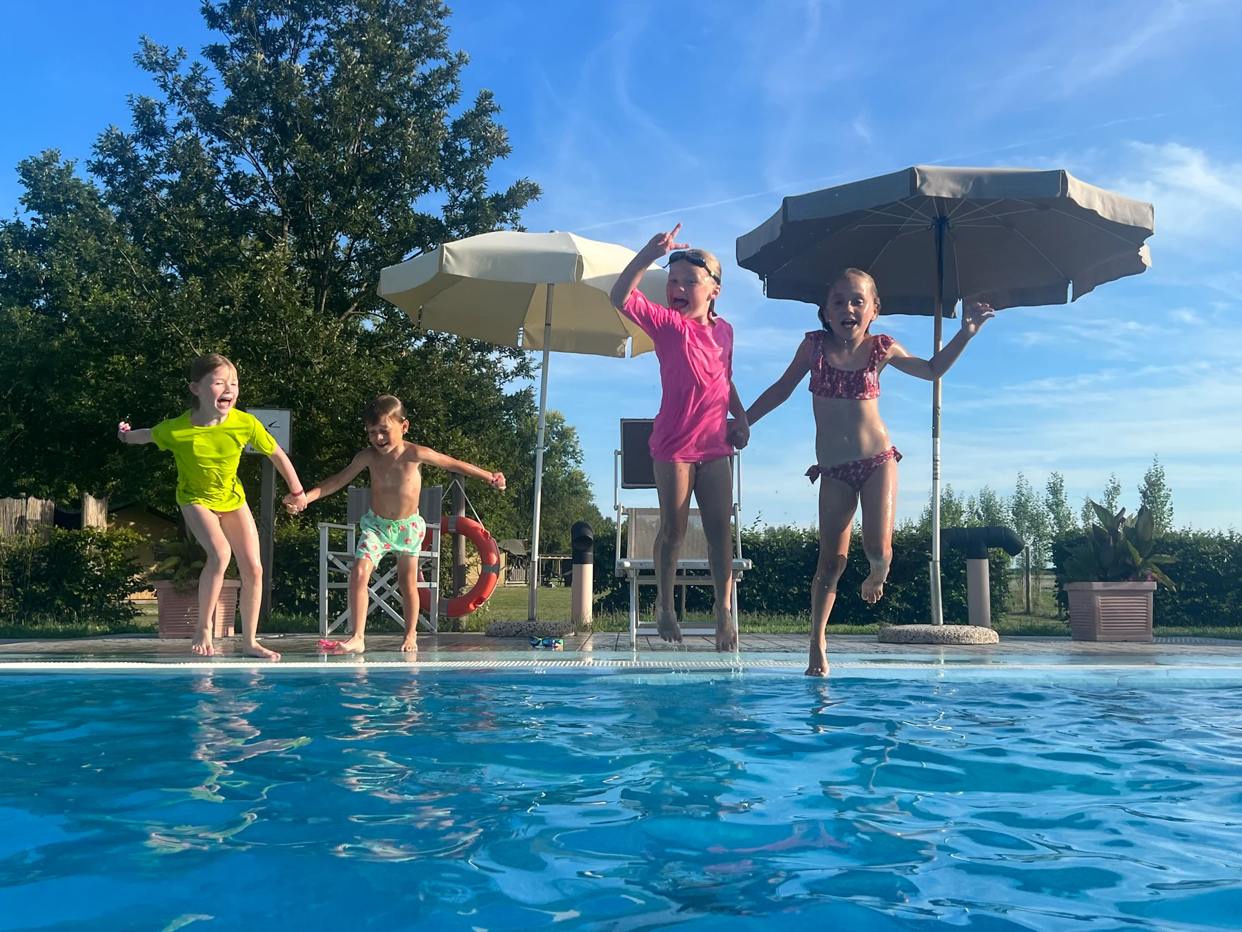 At Agriturismo Tenuta Regina, four kids are leaping into a swimming pool, grasping each other's hands, with umbrellas and foliage in the backdrop.