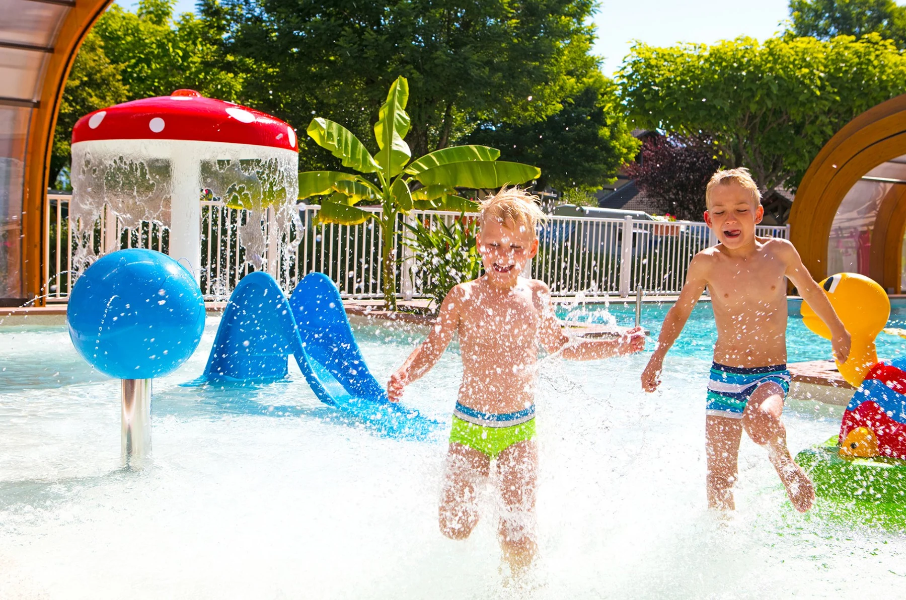 At Le Coin Tranquille, two boys are playing and splashing in a children's pool with water attractions on a bright, sunny day.