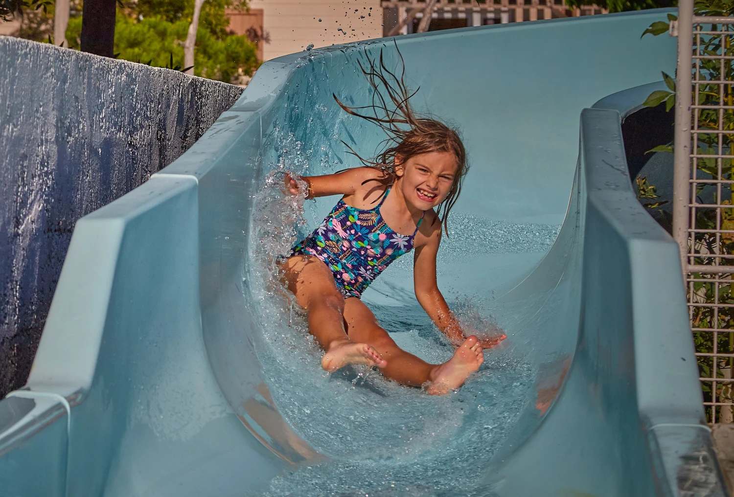 At Crin Blanc, a child glides down a water slide in the open air, splashing joyfully and beaming, encircled by lush greenery and sunlit buildings.