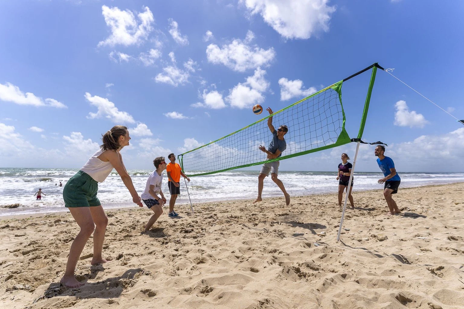 At Amis de la Plage, individuals engage in beach volleyball, striking the ball over the net on a sunny shoreline with waves in the backdrop.