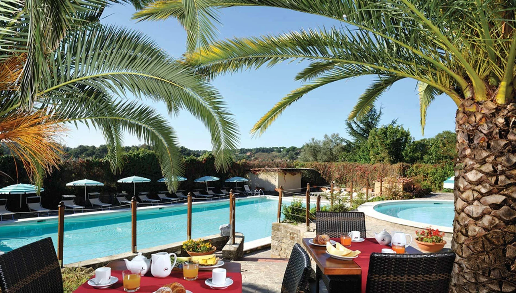 At Happy Village & Camping, a breakfast table arranged for four is situated beside a pool, shaded by umbrellas under palm trees.
