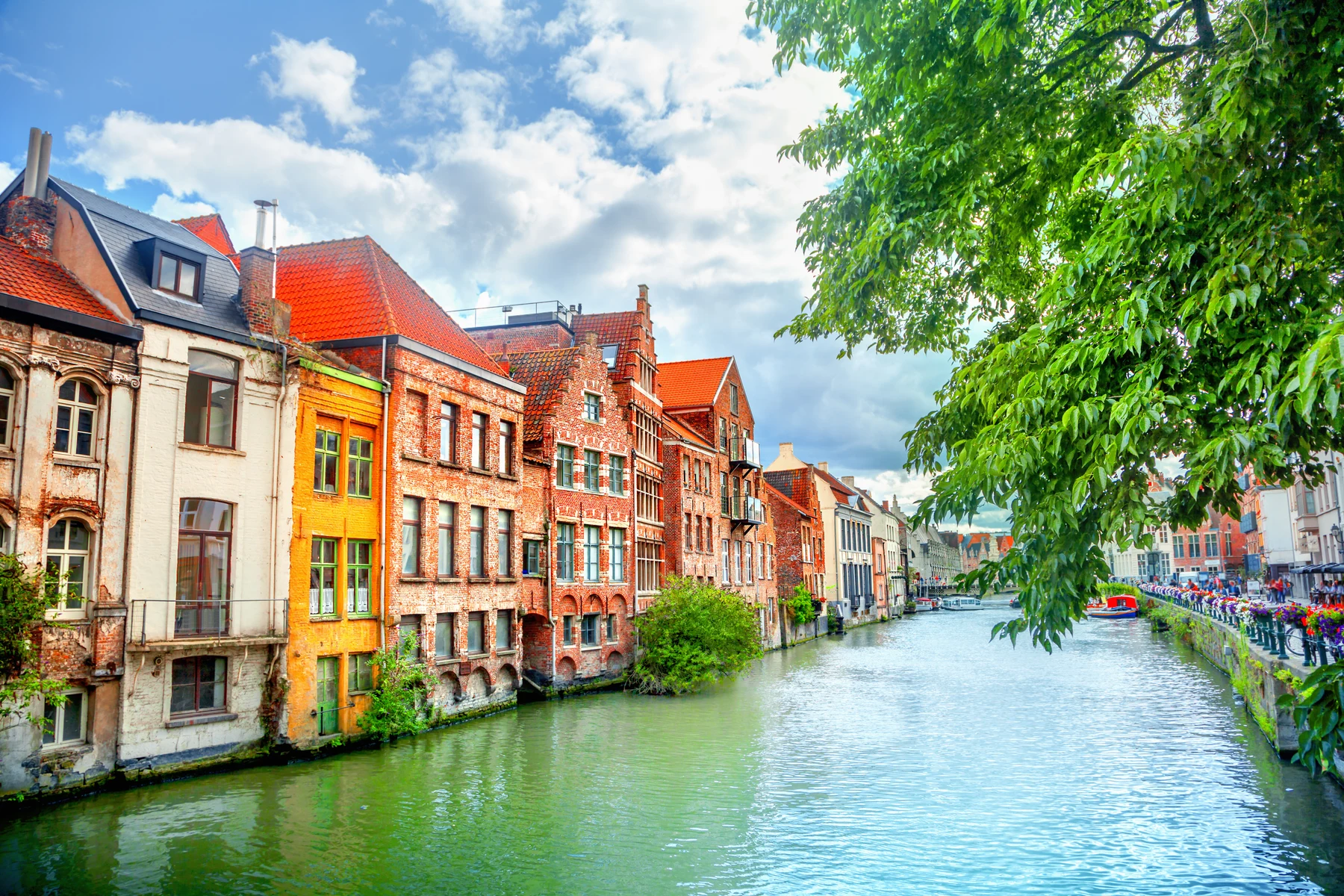 At Urban Gardens Gent, vibrant structures line a serene waterway, bordered by a tree, beneath a partly cloudy sky.