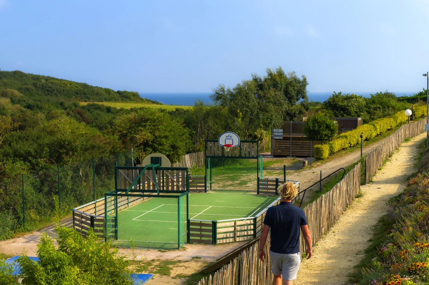 At le Châtelet, an individual strolls along a trail adjacent to an outdoor basketball court encircled by lush vegetation and the ocean in the backdrop.