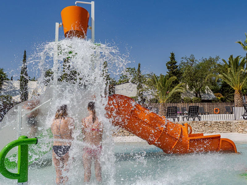 At Orangeraie, two individuals are positioned beneath a large container of water pouring into a swimming area featuring water slides, palm trees, and lounge chairs.