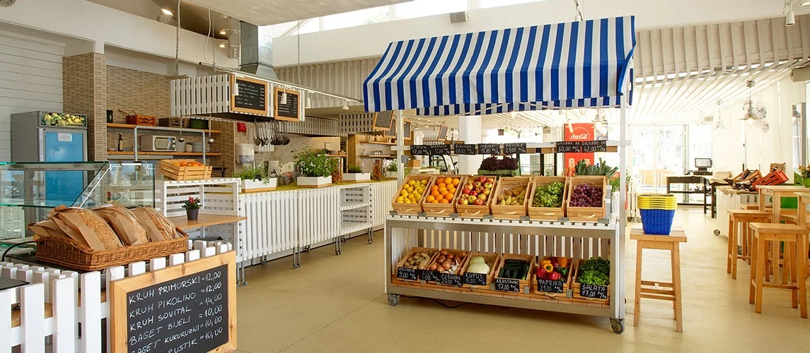 At Bunculuka, a market stall showcases vibrant fruits and vegetables beneath a blue-striped awning within a lively, contemporary marketplace.